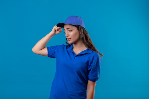 Молодая доставщица в синей униформе и кепке невежественная и смущенная почесывает голову, не имея ответа, стоя на синем фоне