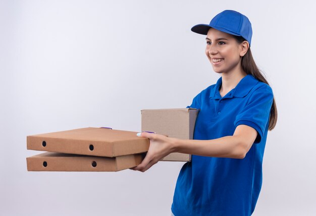 Молодая доставщица в синей форме и кепке дарит покупателю коробки с пиццей, дружелюбно улыбаясь