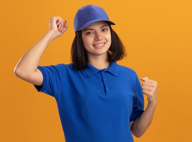 Giovane ragazza delle consegne in uniforme blu e berretto felice ed eccitato che stringe i pugni che rallegrano il suo successo in piedi sopra la parete arancione