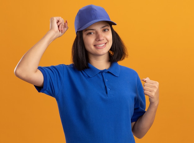 파란색 유니폼과 모자에 젊은 배달 소녀 오렌지 벽 위에 서있는 그녀의 성공을 기뻐하는 행복하고 흥분 떨리는 주먹