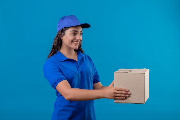 Young delivery girl in blue uniform and cap holding box package giving it to a customer standing over blue background