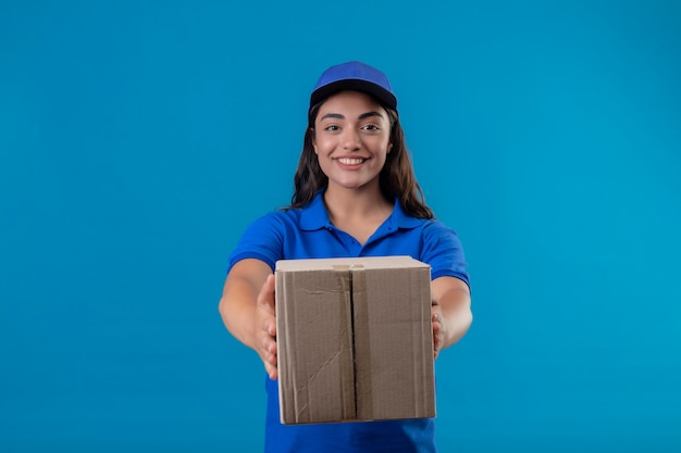 Free photo young delivery girl in blue uniform and cap holding box package giving it to a customer standing over blue background