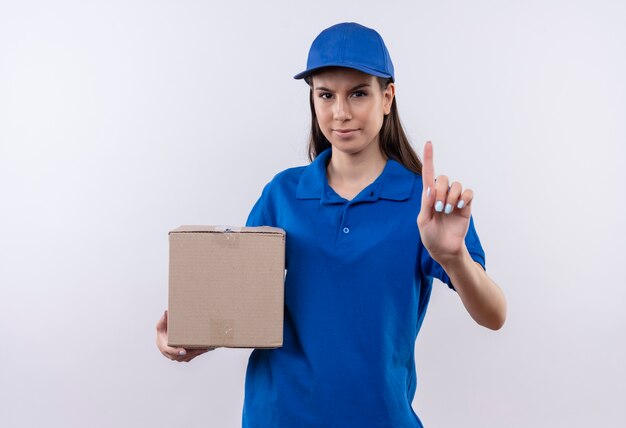 Young delivery girl in blue uniform and cap holding box package showing index finger warning with frowning face 