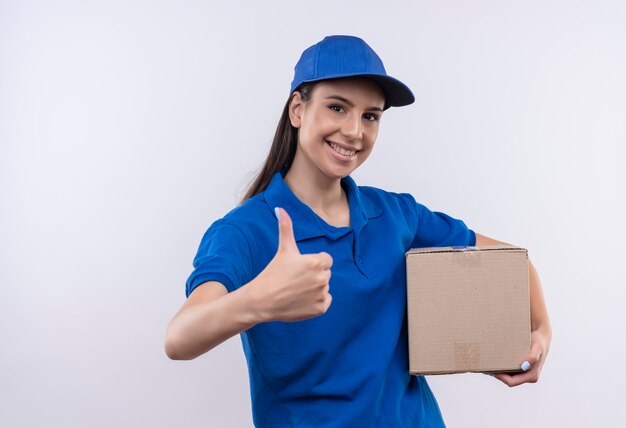 Молодая доставщица в синей униформе и кепке, держащая коробку, уверенно улыбается, показывает палец вверх
