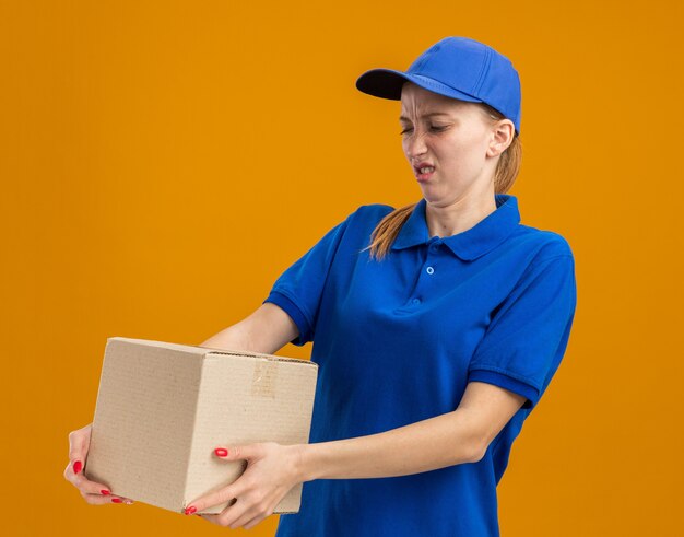 Молодая доставщица в синей форме и кепке держит картонную коробку, глядя на нее, смущенная и недовольная, стоя над оранжевой стеной