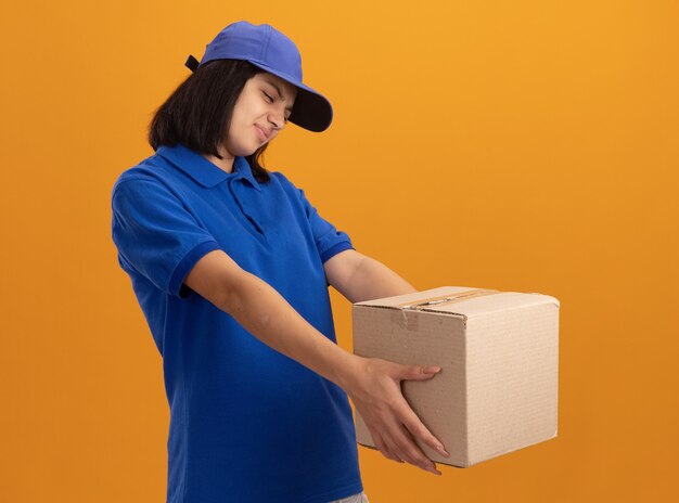 young delivery girl in blue uniform and cap holding cardboard box looking at it with disgusted expression standing over orange wall