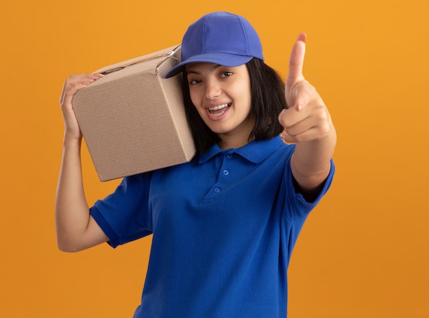 Giovane ragazza di consegna in uniforme blu e cappuccio che tiene la scatola di cartone che sorride allegramente indicando con il dito indice in piedi sopra la parete arancione