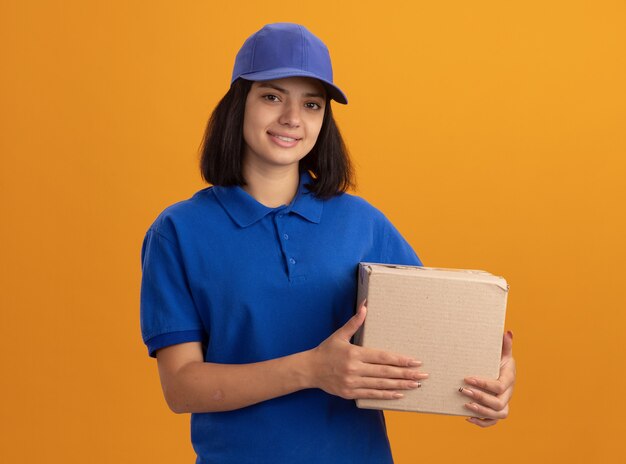 Young delivery girl in blue uniform and cap holding cardboard box  smiling friendly standing over orange wall