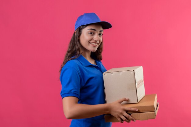 Young delivery girl in blue uniform and cap holding cardboard boxes looking at camera smiling confident happy and positive standing over pink background