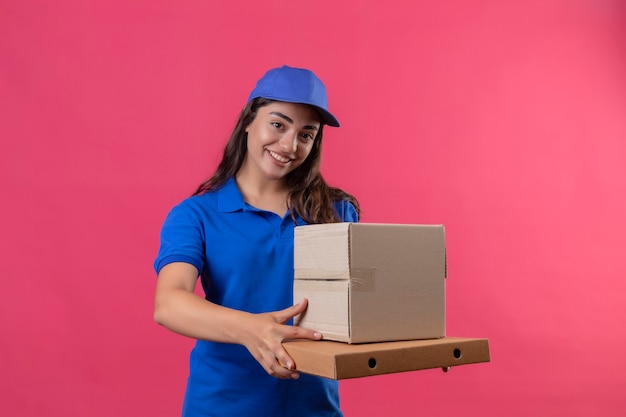 Молодая доставщица в синей форме и кепке держит картонные коробки, глядя в камеру, улыбаясь уверенно, счастливым и позитивным положением на розовом фоне