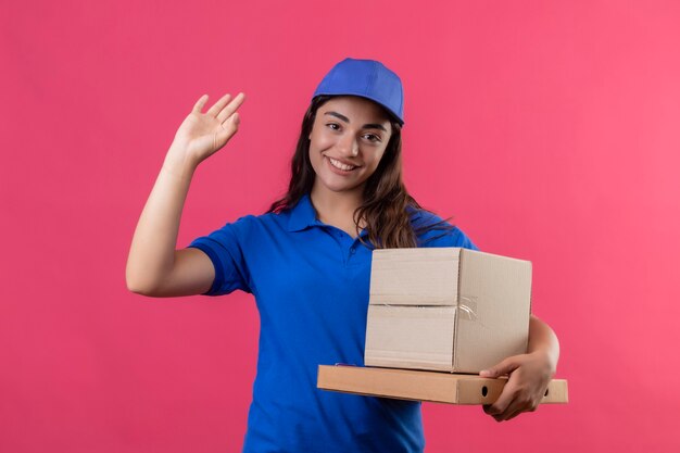 Молодая доставщица в синей форме и кепке держит картонные коробки, глядя в камеру, улыбаясь дружелюбно машет рукой, стоя на розовом фоне