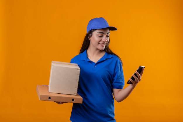 Young delivery girl in blue uniform and cap holding cardboard boxes looking at screen of smartphone smiling confident standing over yellow background