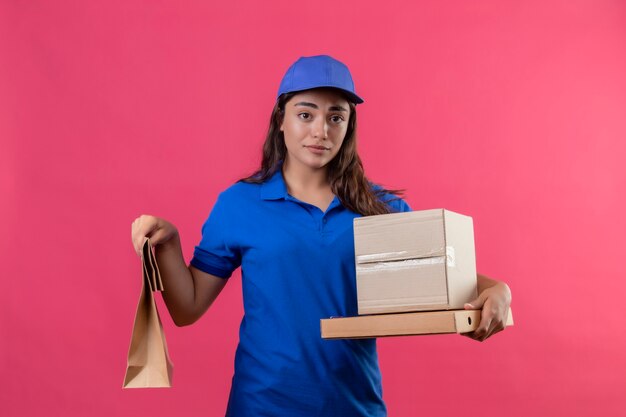 Young delivery girl in blue uniform and cap holding cardboard boxes and paper package looking unhappy standing with sad expression on face over pink background