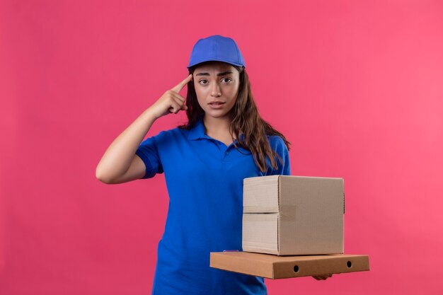 Молодая доставщица в синей форме и кепке держит картонные коробки, указывая на храм за ошибку, выглядит встревоженной, стоя на розовом фоне