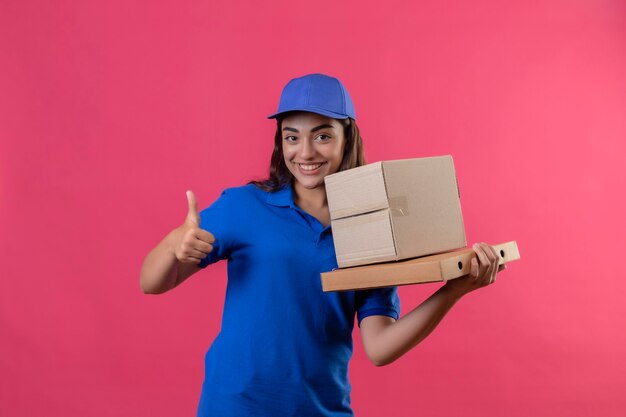 파란색 제복을 입은 젊은 배달 소녀와 분홍색 배경 위에 서있는 엄지 손가락을 유쾌하게 보여주는 골판지 상자를 들고 모자