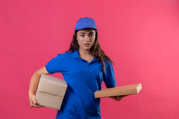 Young delivery girl in blue uniform and cap holding cardboard boxes standing with sad expression on face over pink background