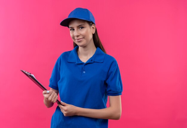 Free photo young delivery girl in blue uniform and cap holding clipboard looking at camera with confident smile on face