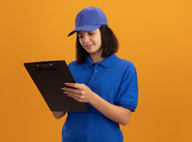 Young delivery girl in blue uniform and cap holding clipboard looking at it with smile on face standing over orange wall