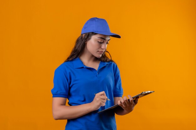 Young delivery girl in blue uniform and cap holding clipboard looking at it writing something with serious face standing over yellow background