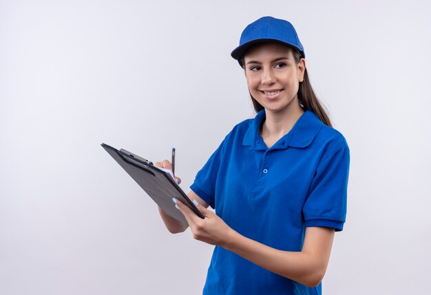 Young delivery girl in blue uniform and cap holding clipboard and pen looking aside with confident smile on face 