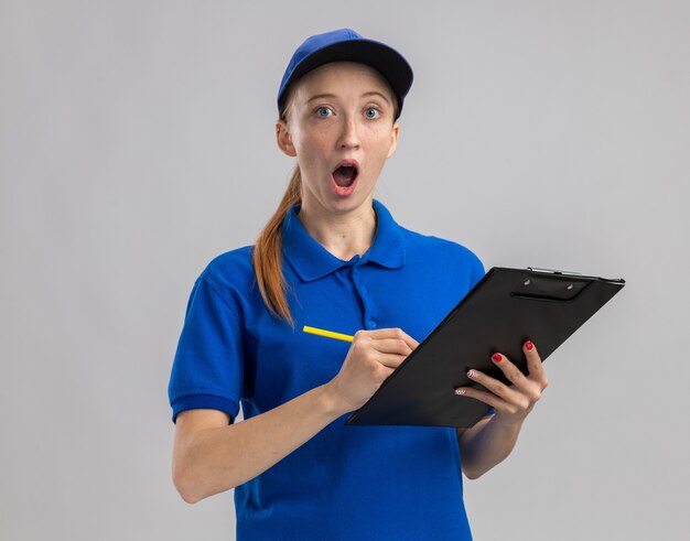 Giovane ragazza delle consegne in uniforme blu e cappello che tiene appunti e matita che sembra sorpresa a scrivere qualcosa in piedi sul muro bianco