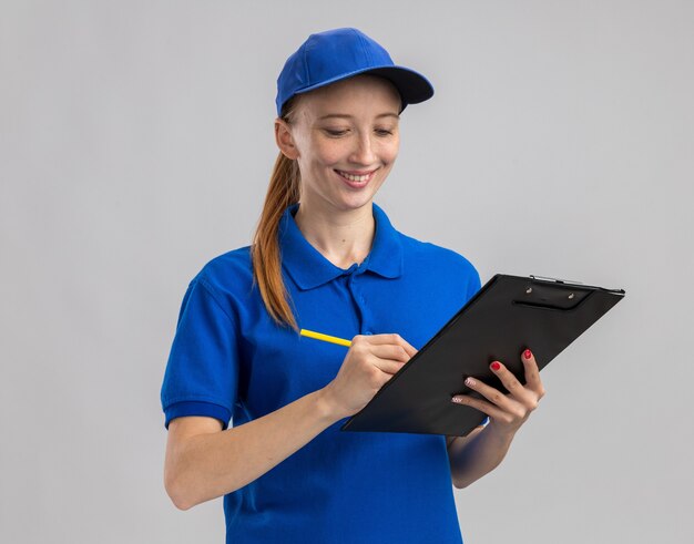 Young delivery girl in blue uniform and cap holding clipboard and pencil smiling confident writing something