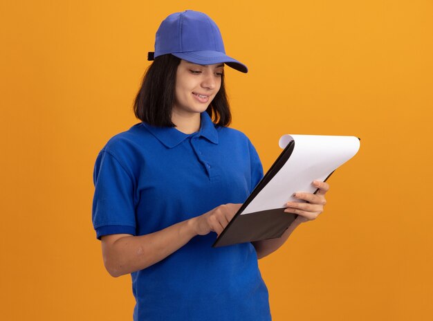 Молодая доставщица в синей форме и кепке держит буфер обмена с пустыми страницами, глядя на буфер обмена, читает и улыбается, стоя над оранжевой стеной