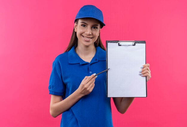 Молодая доставщица в синей форме и кепке держит буфер обмена с пустыми страницами, указывающими на него ручкой с просьбой подписать с улыбкой на лице