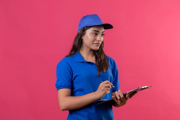 Young delivery girl in blue uniform and cap holding clipboard with pen looking aside with confident smile standing over pink backgground