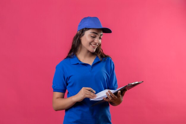 Молодая доставщица в синей форме и кепке держит буфер обмена с ручкой, глядя на нее, уверенно улыбаясь, стоя на розовом фоне