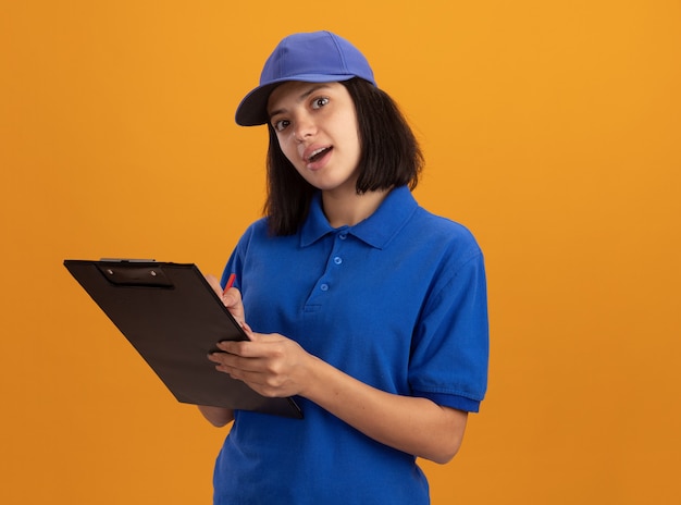 Young delivery girl in blue uniform and cap holding clipboard writing something smiling standing over orange wall