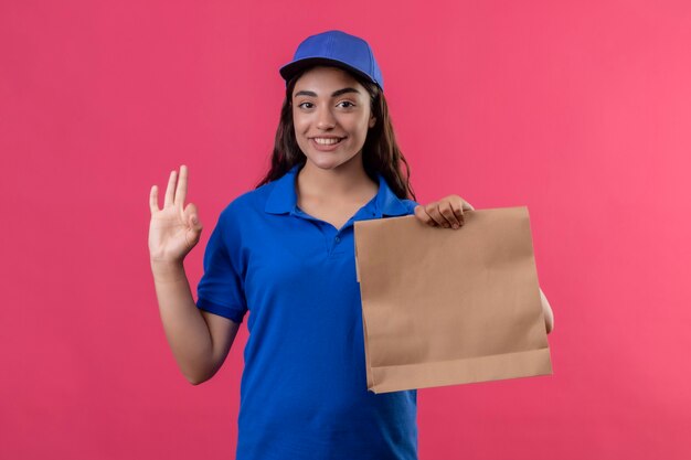 Молодая доставщица в синей форме и кепке держит бумажный пакет, глядя в камеру, улыбаясь дружелюбно, делая хорошо, знак, стоящий на розовом фоне