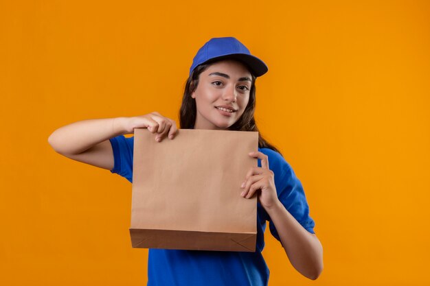 Young delivery girl in blue uniform and cap holding paper package looking at camera smiling friendly standing over yellow background