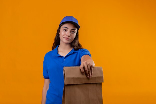 Giovane ragazza delle consegne in uniforme blu e cappuccio che tiene il pacchetto di carta che guarda l'obbiettivo con un sorriso fiducioso sul viso in piedi su sfondo giallo