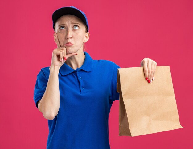 Giovane ragazza delle consegne in uniforme blu e berretto con in mano un pacchetto di carta che guarda perplesso in piedi sul muro rosa pink