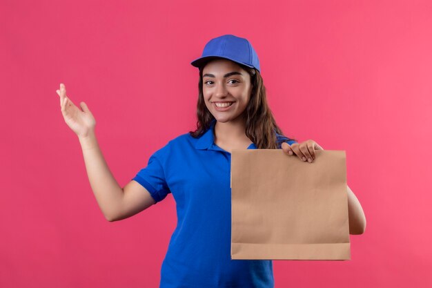 Молодая доставщица в синей форме и кепке, держащая бумажный пакет, представляющая рукой, смотрящую в камеру с улыбкой на лице, счастливым и позитивным положением на розовом фоне