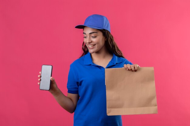 Young delivery girl in blue uniform and cap holding paper package showing smartphone looking at it smiling cheerfully standing over pink background