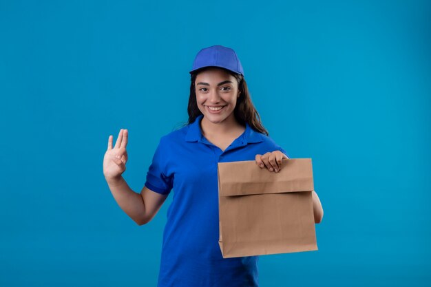 파란색 제복을 입은 젊은 배달 소녀와 파란색 배경 위에 서있는 확인 서명을 유쾌하게 웃고있는 종이 패키지를 들고 모자