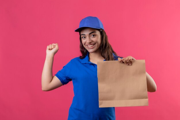 青い制服を着た若い配達の少女と彼女の成功と喜びピンクの背景の上に立って勝利を喜んで元気に拳を上げる笑顔の紙パッケージを保持しているキャップ