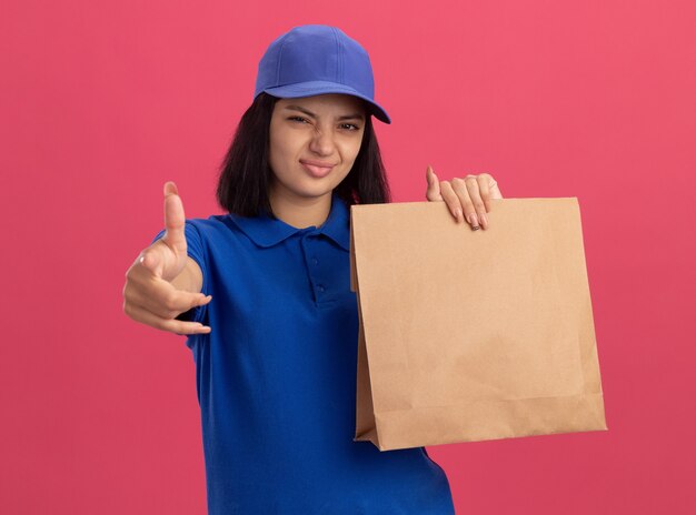 Молодая доставщица в синей униформе и кепке держит бумажный пакет с уверенным выражением лица, указывая указательным пальцем на вас, стоя над розовой стеной