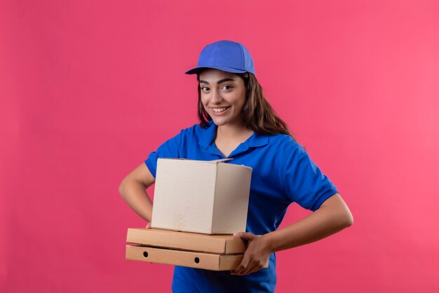 Молодая доставщица в синей униформе и кепке держит коробки для пиццы и коробку, глядя в камеру, весело улыбаясь и радостно улыбаясь на розовом фоне