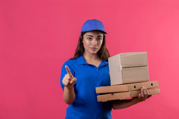 Молодая доставщица в синей форме и кепке держит коробки для пиццы и коробку, указывая пальцем на что-то, смотрящее в камеру с уверенным выражением лица, стоящее на розовом фоне