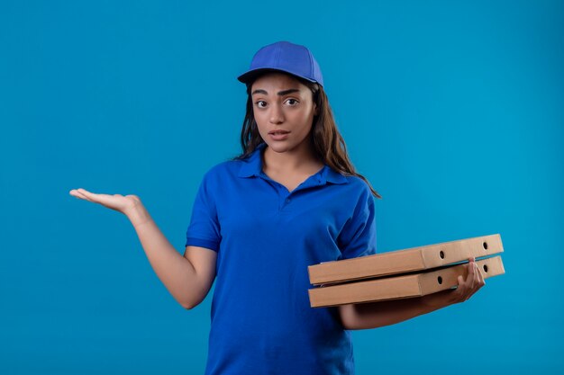 Young delivery girl in blue uniform and cap holding pizza boxes clueless and confused standing with arm raised having no answer over blue background