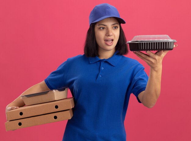 ピンクの壁の上に立って驚いて驚いたパッケージを見てピザの箱と食品パッケージを保持している青い制服と帽子の若い配達の女の子