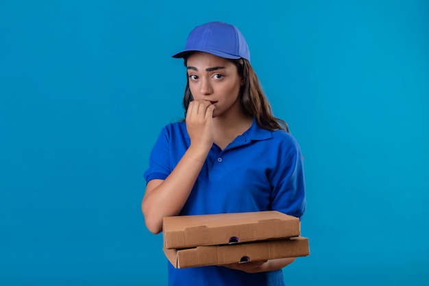 Young delivery girl in blue uniform and cap holding pizza boxes looking at camera nervous and stressed biting nails standing over blue background