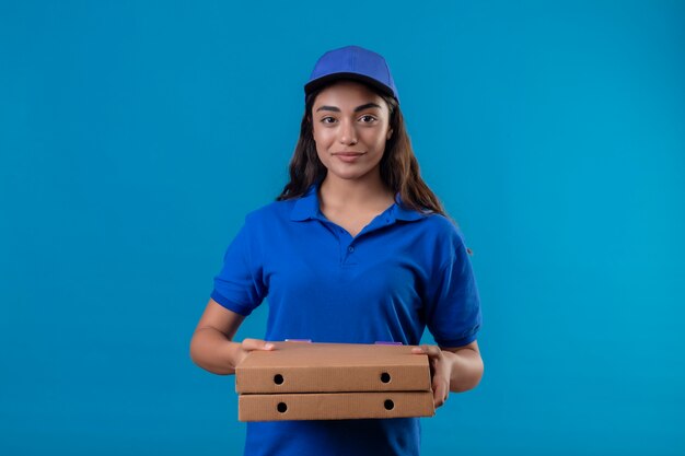 青い制服を着た若い配達の少女とカメラを見てピザの箱を持ってキャップ青い背景に自信を持って幸せと肯定的な立っている笑顔