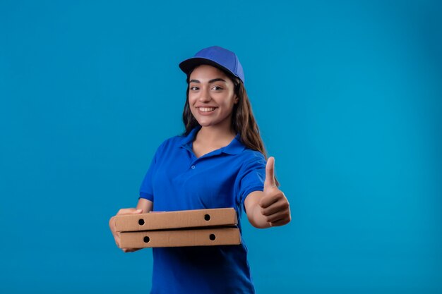 Молодая доставщица в синей униформе и кепке держит коробки для пиццы, глядя в камеру, улыбаясь дружелюбно, счастливо и позитивно, показывая большие пальцы руки вверх, стоя на синем фоне