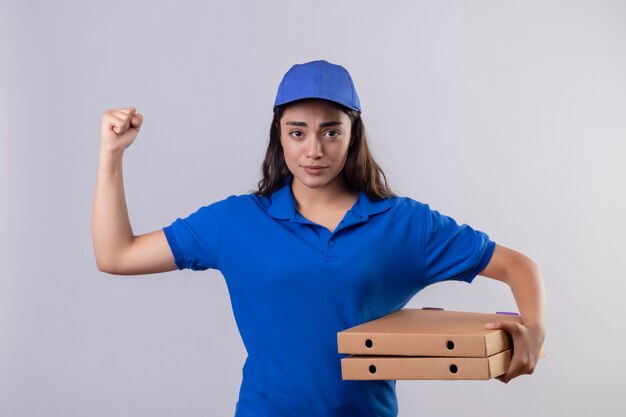 Молодая доставщица в синей форме и кепке держит коробки для пиццы, поднимая кулак, глядя в камеру с хмурым лицом, стоящим на белом фоне