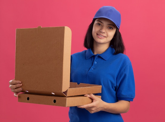 Молодая доставщица в синей форме и кепке держит коробки для пиццы, дружелюбно улыбаясь, стоя у розовой стены