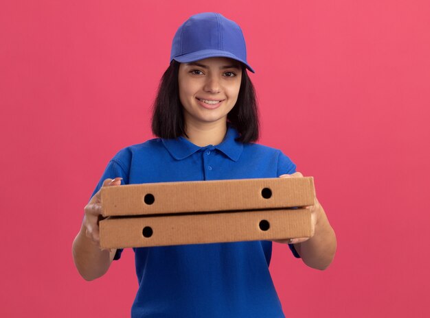 ピンクの壁の上に立っている幸せそうな顔で笑顔のピザの箱を保持している青い制服と帽子の若い配達の女の子
